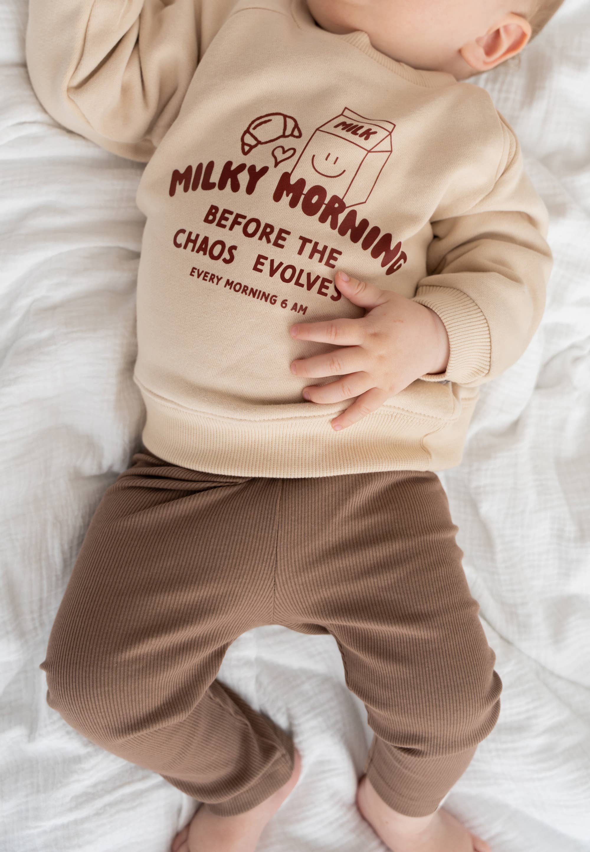 Baby draagt beige Liliput sweater met “Milky Morning” tekst en print