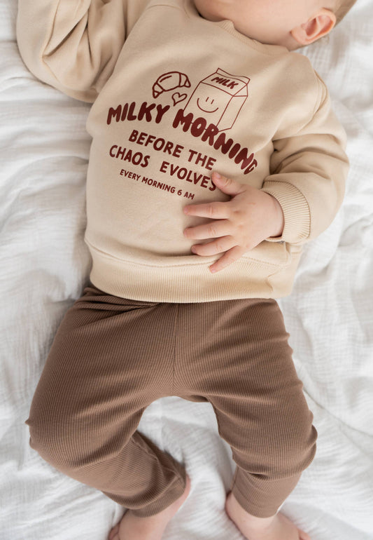 Baby draagt beige Liliput sweater met “Milky Morning” tekst en print